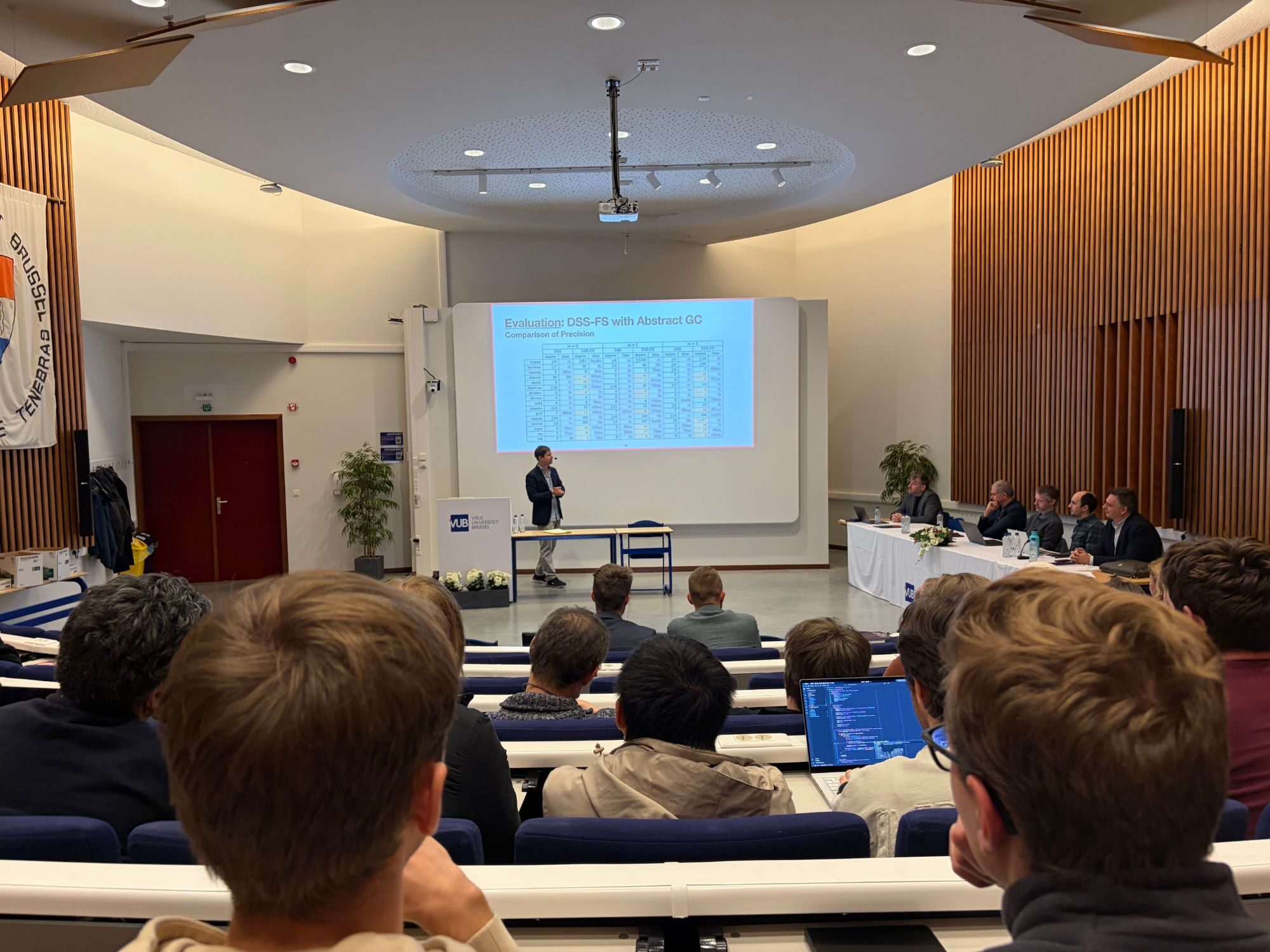 Noah Van Es presenting his PhD defence in a VUB auditorium, seen from the audience, with the jury seated at a table to the right
